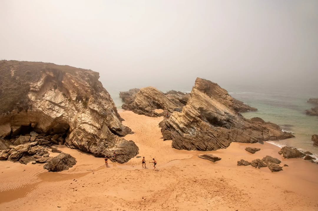 Praia entre rochedos envolta em bruma, com figuras a brincar na areia