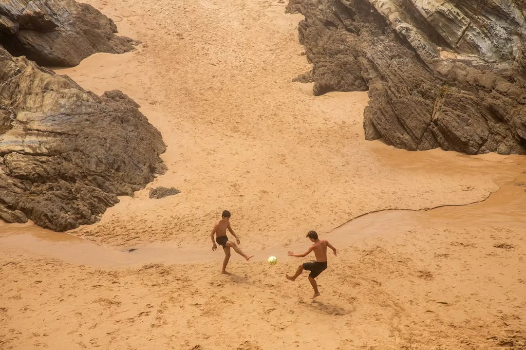 Dois rapazes jogam futebol na areia molhada entre rochedos
